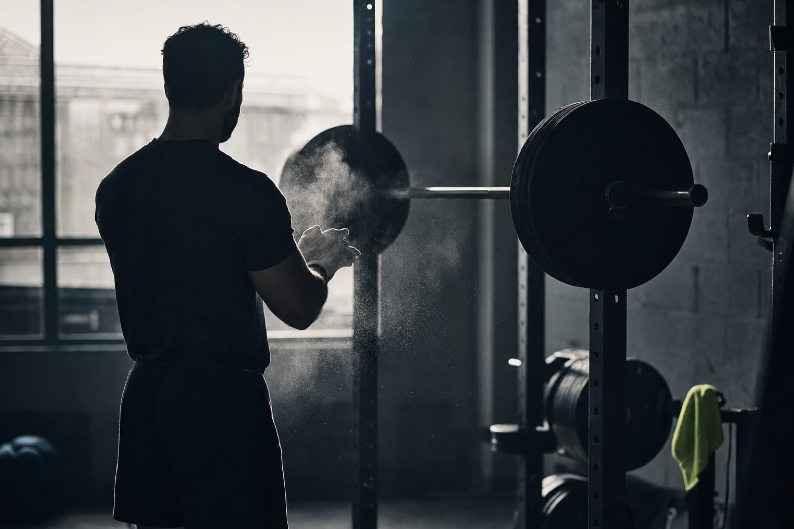 A lifter chalking their hands at a squat rack, backlit by a gym window
